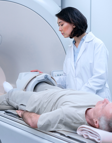Un paciente está acostado esperando una tomografía computarizada mientras un técnico de radiología está de pie junto a la mesa deslizante